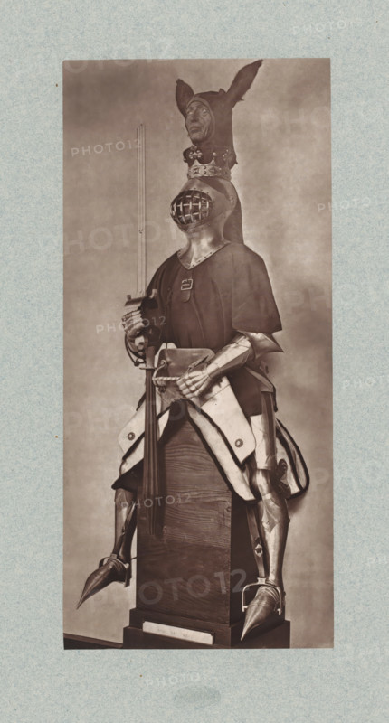15th-century armor for a cavalryman from the army of Charles VII of France, from the collection of the Musée d'Artillerie in Paris, photomechanical print, This print is part of a cover with 36 prints, a title page and a list of images., check stamp: 'PHOTOGLYPTIE, LEMERCIER ET Cie, PARIS', Paris, cliché maker: Lemercier & Cie., c. 1877 - in or before 1882, Lemercier & Cie., photographer, cliché maker, paper, Woodburytype, Woodburytypie, height 268 mm x width 122 mm, armour, A monochrome photograph shows a full suit of armor displayed upright on a wooden pedestal, with a helmet featuring a grilled visor and a separate decorative headpiece resembling an animal head with long pointed ears placed above it, the armor including articulated gauntlets, a breastplate partially covered by a dark surcoat, and metal greaves and sabatons with pointed toes; the figure holds a long polearm with a hand on its haft and has a cloth or padded skirt draped over the hips, all set against a plain studio bac...