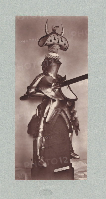 15th-century armor for a cavalryman from the army of Charles VII of France, from the collection of the Musée d'Artillerie in Paris, photomechanical print, This print is part of a cover with 36 prints, a title page and a list of images., check stamp: 'PHOTOGLYPTIE, LEMERCIER ET Cie, PARIS', Paris, cliché maker: Lemercier & Cie., c. 1877 - in or before 1882, Lemercier & Cie., photographer, cliché maker, paper, Woodburytype, Woodburytypie, height 266 mm x width 118 mm, armour, A full suit of polished plate armor displayed on a wooden stand, shown from a side angle with a lance held horizontally through a rounded shoulder plate, a rounded closed helmet with a decorative wide-brimmed crest topped by a small sculpted animal and horn-like projections, a cloth-lined collar visible beneath the helmet, articulated pauldrons, a segmented fauld and tassets over the hips, articulated cuisses and greaves on the legs, metal sabatons on the feet, and ornamental fluting and rivets on several pieces; th...