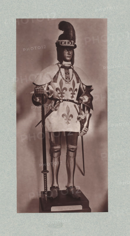 15th-century armor with various thrusting weapons from the army of Louis XI of France, from the collection of the Musée d'Artillerie in Paris, photomechanical print, This print is part of a cover with 36 prints, a title page and a list of images., check stamp: 'PHOTOGLYPTIE, LEMERCIER ET Cie, PARIS', Paris, cliché maker: Lemercier & Cie., c. 1877 - in or before 1882, Lemercier & Cie., photographer, cliché maker, paper, Woodburytype, Woodburytypie, height 278 mm x width 129 mm, hacking and thrusting weapons: sword, armour, A sepia-toned full-length photograph of a mannequin or statue dressed in medieval-style armor and a decorative surcoat, standing on a wooden plinth; the figure wears a helmet with a tall feather plume and a jeweled band, a layered shoulder cape over the surcoat, and a chain around the neck, with stylized fleur-de-lis motifs visible on the front of the surcoat; the arms show articulated plate armor and gauntlets, one hand resting on a long pole weapon and the other hol...