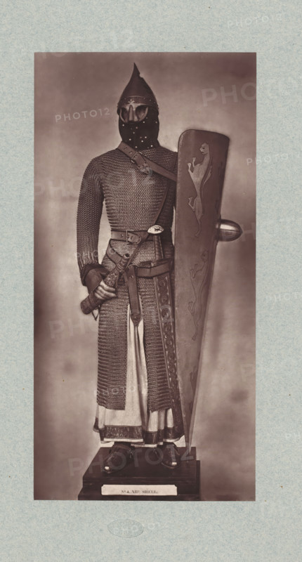 12th-century armor from the army of Louis VI of France, from the collection of the Musée d'Artillerie in Paris, photomechanical print, This print is part of a cover with 36 prints, a title page and a list of images., check stamp: 'PHOTOGLYPTIE, LEMERCIER ET Cie, PARIS', Paris, cliché maker: Lemercier & Cie., c. 1877 - in or before 1882, Lemercier & Cie., photographer, cliché maker, paper, Woodburytype, Woodburytypie, height 275 mm x width 135 mm, armour, helved weapons, polearms (for striking, hacking, thrusting): lance, A full-length sepia-toned photograph shows a person standing on a pedestal wearing medieval-style armor including a conical helmet with a pointed top and a face-covering mask with eye openings, a long chainmail hauberk extending down over a tunic, and gauntleted hands; a broad belt and cross-body strap with decorative buckles secure a sheathed sword at the waist, and one hand grasps the sword hilt while the other rests behind a tall kite-shaped shield decorated with st...