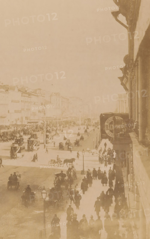 The Nevsky Perspective, View of Nevsky Prospekt in Saint Petersburg, Russia, Saint-Pétersbourg, photograph, page, Saint Petersburg, 1890-06 - 1890-07, photographer, Leningradskaya, paper, albumen print, height 164 mm x width 222 mm, street, Nevsky Prospekt, A faded rectangular photograph mounted on aged paper with decorative printed borders, showing an upside-down sepia street scene with people, horse-drawn carriages, and buildings, framed by a thin red inner line and ornate brown corner embellishments, with scattered spots and discoloration on the surrounding beige background.