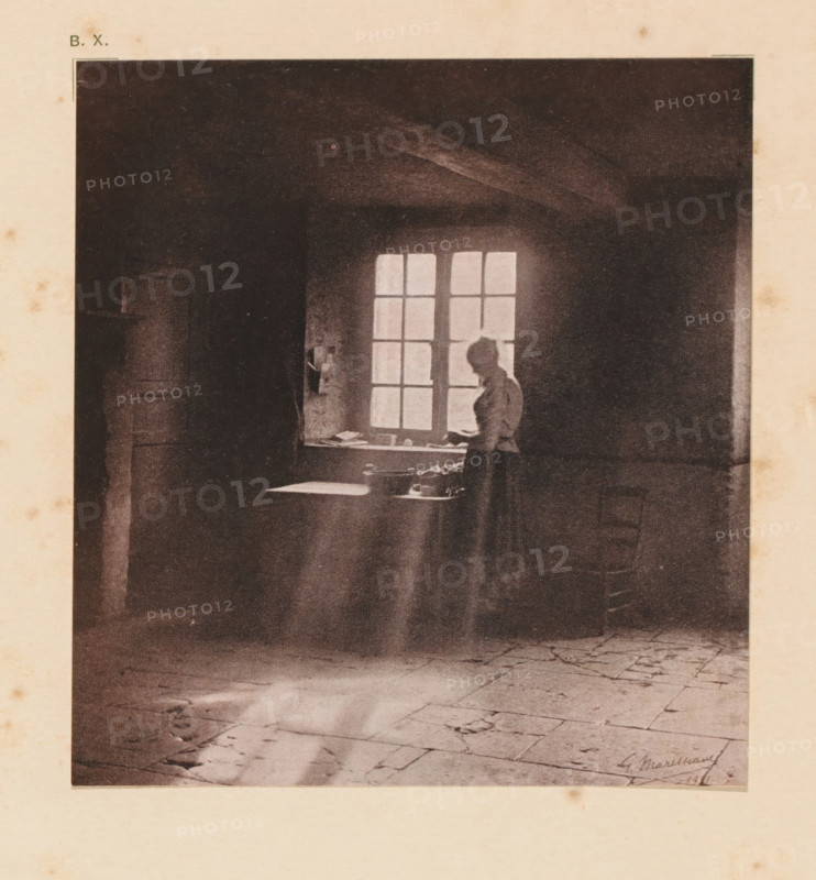 Woman at the window, page, photomechanical print, inscription: 'Interior by G. Marissiaux', inscription: 'BX', photographer: Gustave Marissiaux, cliché maker: anonymous, c. 1900 - in or before 1905, paper, two-color autotype, height 132 mm x width 123 mm, adult woman, interior of the house, A dark interior scene shows a woman standing by a multi-pane window with light streaming through, a table beneath the window holding small objects, a high ceiling with visible beams, a tiled floor with cracks and worn areas, and strong contrasts between illuminated and shadowed areas.