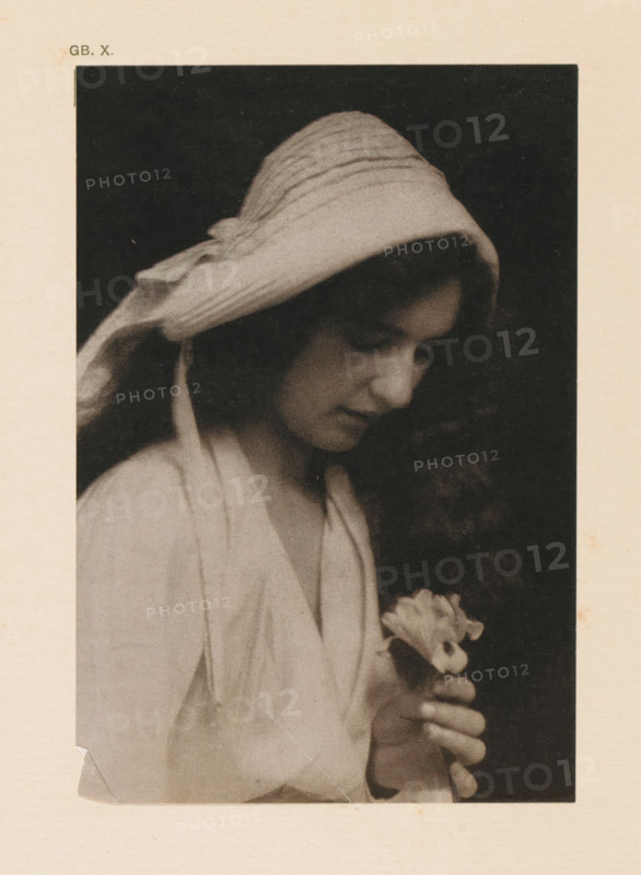 Portrait of an unknown woman with an iris, page, photomechanical print, inscription: 'GB. X.', inscription: 'The Iris by CF Juston', photographer: CF Juston, cliché maker: anonymous, c. 1900 - in or before 1905, paper, two-color autotype, height 149 mm x width 101 mm, historical persons - BB - woman, flowers: iris, A sepia-toned portrait of a young woman shown in profile against a dark background, wearing a textured headscarf that drapes over her hair and shoulders, her eyes cast downward and her expression neutral, her long curly hair visible beneath the scarf, a light-colored loose garment covering her upper body, and her hands gently holding a small cluster of light flowers near her chest.