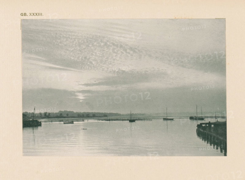 Water landscape with sailing boats, page, photomechanical print, inscription: 'Land & sea by WJ Day', inscription: 'GB. XXXIII.', photographer: William James Day, cliché maker: anonymous, c. 1900 - in or before 1905, paper, autotypie, height 97 mm x width 144 mm, sailing-ship, sailing-boat, landscapes with waters, waterscapes, seascapes (in the temperate zone), A grayscale scene showing a calm body of water with reflections, small boats moored or floating, and vertical masts rising from some of the boats, a distant shoreline with low tree silhouettes, a pier or jetty extending into the water on the right with visible posts and railings, and a sky filled with layered clouds and streaks of light near the horizon.