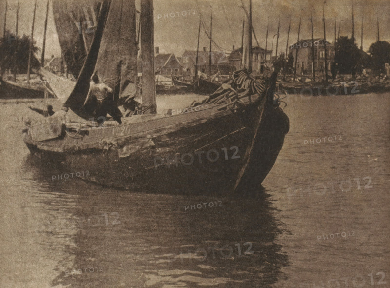 Sailboat setting sail with the sail being hoisted, page, photomechanical print, inscription: 'GB. XXXIV.', inscription: 'Hauling sail by Arthur Marshall', photographer: Arthur Marshall, cliché maker: anonymous, c. 1900 - in or before 1905, paper, autotype, height 114 mm x width 154 mm, sailing-ship, sailing-boat (view, vehicle, ship, etc.), A sepia-toned photograph showing a close view of a wooden sailboat floating on calm water with its bow angled toward the viewer, a coiled sail and ropes piled on the deck near the bow, a partially raised sail and mast amidships, figures on the boat working with the rigging, reflections of the boat and masts in the water, and a distant shoreline lined with more masts and several buildings silhouetted against the sky.