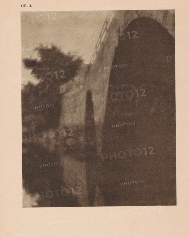 View of a bridge at Ipswich, Massachusetts, page, photomechanical print, inscription: 'US. II.', inscription: 'The bridge Ipswich by Alvin L Coburn', photographer: Alvin Langdon Coburn, Ipswich (Massachusetts), cliché maker: anonymous, c. 1900 - in or before 1905, North America, paper, autotypie, height 145 mm x width 115 mm, bridge in city across river, canal, A grainy sepia-toned photograph showing a large archway on the right with a shadowed interior and a textured wall leading away from it, trees with dark, blurred foliage on the left, a railing or fence running along the top of the wall, and an indistinct foreground of blurred shapes and tones.