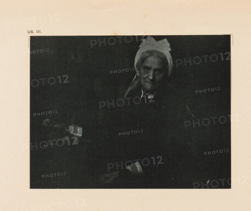 Portrait of an unknown old woman, page, photomechanical print, inscription: 'Portrait of an old lady by Gertrude Käsebier', inscription: 'US. III.', photographer: Gertrude Käsebier, cliché maker: anonymous, c. 1900 - in or before 1905, paper, autotypie, height 110 mm x width 144 mm, historical persons - BB - woman, old woman, A dark, rectangular photograph centered on a pale textured background shows an older woman wearing a light-colored head covering with a bow-like shape at the top and a dark garment, her face partially illuminated against a black backdrop, with one hand visible near the bottom holding a small striped object.