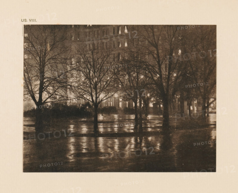 Street view at night, page, photomechanical print, inscription: 'US. VIII.', inscription: 'Reflections night by Alfred Stieglitz', photographer: Alfred Stieglitz, cliché maker: anonymous, c. 1900 - in or before 1905, paper, two-color autotype, height 109 mm x width 142 mm, street, night, A dark, grainy scene shows several leafless trees with bare branches silhouetted against a softly illuminated background, their trunks reflected in a wet, glistening surface below; behind the trees faint, blurred shapes of a multi-story building appear with small bright rectangles of light, and the overall image has a muted, sepia-toned appearance.
