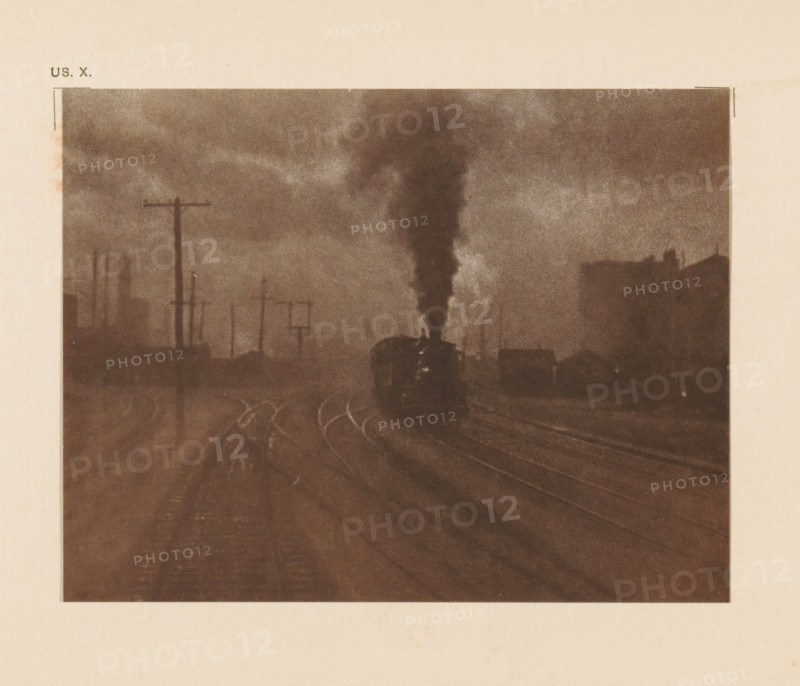 Railway lines with steam train, page, photomechanical print, inscription: 'The hand of man by Alfred Stieglitz', inscription: 'US. X.', photographer: Alfred Stieglitz, cliché maker: anonymous, c. 1900 - in or before 1905, paper, two-color autotype, height 109 mm x width 142 mm, railway, train, A sepia-toned image showing a railway scene with tracks curving into the distance, a dark locomotive near the center emitting a tall plume of smoke that rises into a cloudy sky, silhouettes of telegraph poles and industrial structures along the left side, and faint outlines of buildings on the right, all framed by a pale border.