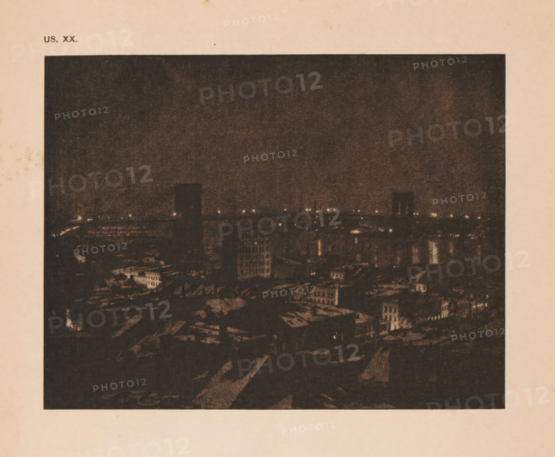 View of New York with the Brooklyn Bridge, page, photomechanical print, inscription: 'US. XX.', inscription: 'The bridge by JF Strauss', photographer: John Francis Strauss, cliché maker: anonymous, c. 1900 - in or before 1905, paper, autotypie, height 112 mm x width 146 mm, city-view in general, 'veduta', bridge in city across river, canal, North America, New York (city), Brooklyn Bridge, A dark, grainy nighttime cityscape framed by a light border, showing a skyline with dimly lit buildings and a river reflecting scattered points of light, with several faintly illuminated structures rising above lower rooftops and a mostly dark sky occupying the upper area.
