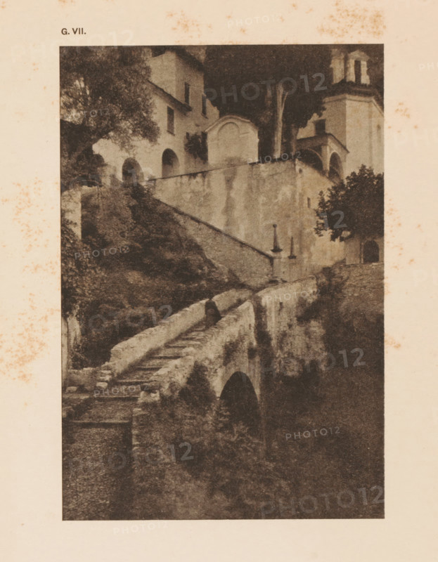 Cityscape in the mountains, page, photomechanical print, inscription: 'Romantic Landscape by Dr. H. Hoek', inscription: 'G. VII.', photographer: H. Hoek, cliché maker: anonymous, c. 1900 - in or before 1905, paper, autotypie, height 148 mm x width 101 mm, city-view in general, 'veduta', A sepia-toned photographic print shows a stone stairway and retaining walls leading up to a cluster of buildings with arched openings and a bell tower. Trees and shrubs grow around the structures, with foliage visible near the building facades and along the stair path. The stone surfaces display weathering and stains, and a narrow arched passage is visible beneath part of the walkway. The image is bordered by a pale margin that has scattered brown spots.