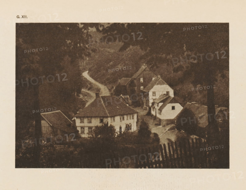 Village view in the Harz Mountains, page, photomechanical print, inscription: 'G. XII.', inscription: 'A Hartz village by HW Müller', photographer: HW Müller, Harz, cliché maker: anonymous, c. 1900 - in or before 1905, paper, autotypie, height 103 mm x width 151 mm, village, mountains, A sepia-toned image of a village scene framed by dark foliage at the top and sides, showing several multi-story houses with sloped roofs and visible windows clustered along a winding road, a small white building with a dark roof set slightly apart, trees and bushes surrounding the buildings, and a foreground area with a fence-like structure appearing as vertical slats.