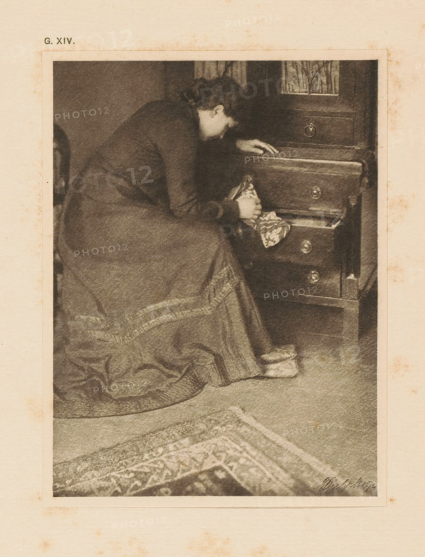 Woman at a chest of drawers, page, photomechanical print, inscription: 'G. XIV.', inscription: 'Memories by R. Rührkoop', photographer: Rudolf Dührkoop, cliché maker: anonymous, c. 1900 - in or before 1905, paper, photoaquatint, height 142 mm x width 106 mm, adult woman, wardrobe, A sepia-toned image of a woman seated beside a wooden chest of drawers, leaning forward with one hand resting on an upper drawer and the other holding a patterned piece of cloth from an open drawer; she wears a long dress with a textured hem and slippers on her feet, and a patterned rug is visible on the floor in front of her.