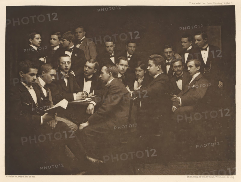 Group of men in suits, including one with a book in hand, page, photomechanical print, photographer: Wilhelm Weimer, Darmstadt, cliché maker: Blechinger & Leykauf, Vienna, c. 1895 - in or before 1900, printer (person), Lower Austria, Hessen, paper, photoaquatint, height 145 mm x width 196 mm, book, adult man (more than eight persons), ensemble of pieces of clothing (men's clothes), A sepia-toned group portrait shows many young men arranged closely together in formal attire, most wearing dark jackets with white shirts and bow ties, seated and standing around a central table; several men hold papers or books, one man looks down at an open book in his hands while another leans forward with hands clasped on the table, faces display neutral expressions, hairstyles are short and neatly combed, a few wear jackets with visible lapels and pocket squares, one man in profile rests an arm on a chair back, another stands with a hand on a seated shoulder, the background is dark and plain, and the im...