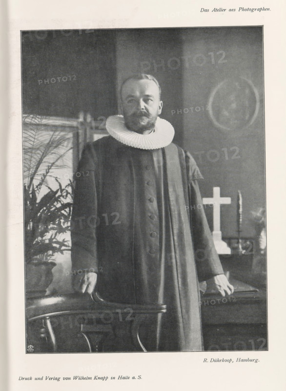 Portrait of an unknown man with collar, page, photomechanical print, inscription: 'Das Atelier aes Photographen.', monogram: 'LEIPZIG, S G', photographer: Rudolf Dührkoop, Hamburg (city), cliché maker: unknown, Leipzig, printer (person): Wilhelm Knapp, Halle an der Saale, c. 1895 - in or before 1900, printer (person), Saxony-Anhalt, Saxony, paper, autotypie, height 201 mm x width 150 mm, historical persons, neck-gear: collar, A black and white portrait of a man standing indoors wearing a dark long robe with buttons down the front and a large pleated white ruff collar, his hands resting on the back of a wooden chair, a potted plant visible behind him to one side, a cross and candlestick on a table in the background, and a circular framed image on the wall.