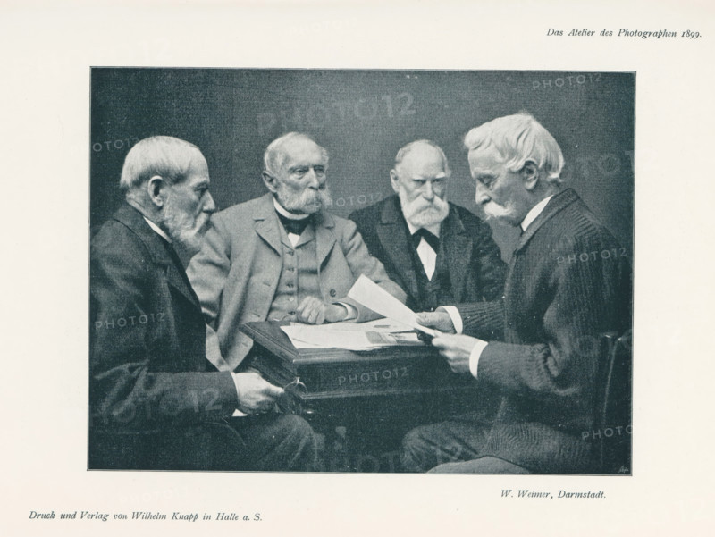 Four men at a table, page, photomechanical print, inscription: 'Das Atelier des Photographen.', photographer: V. de Montano, cliché maker: unknown, printer (person): Wilhelm Knapp, Halle an der Saale, c. 1895 - in or before 1900, printer (person), Saxony-Anhalt, paper, autotypie, height 133 mm x width 180 mm, Print on third unnumbered page after page 124., adult man (four persons), A rectangular black-and-white photograph framed by a light border shows four elderly men seated around a small table holding and looking at papers; all wear suits with jackets and collared shirts, and three have prominent mustaches and beards while one has a mustache and side-parted hair. The man on the far left is shown in profile facing inward, the second man from the left faces slightly toward the camera with folded hands resting on the table, the third man looks down with a stern expression and hands near the papers, and the man on the right holds a sheet of paper and looks at it intently. The table has ...