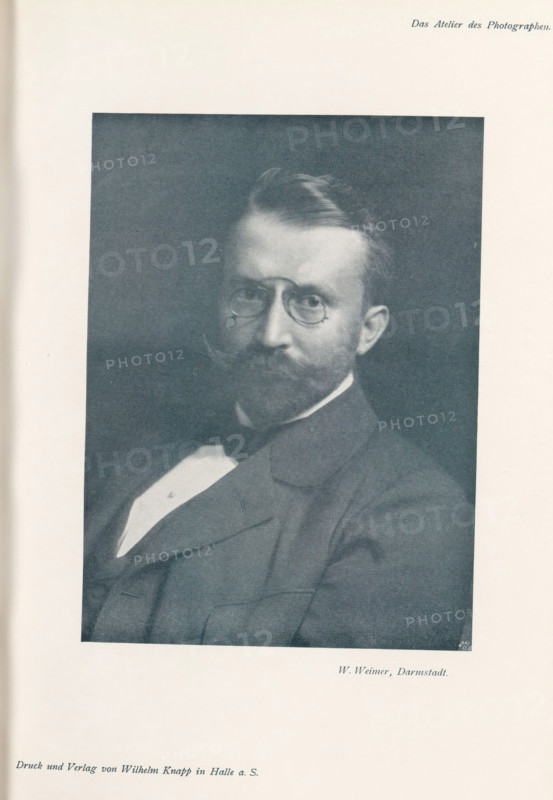 Portrait of an unknown man, page, photomechanical print, inscription: 'Das Atelier des Photographen.', photographer: Wilhelm Weimer, Darmstadt, cliché maker: Meisenbach, Riffarth & Cie., Berlin, printer (person): Wilhelm Knapp, Halle an der Saale, c. 1895 - in or before 1900, printer (person), Hessen, Saxony-Anhalt, paper, autotypie, height 167 mm x width 123 mm, Print on third unnumbered page after page 164., historical persons, A monochrome portrait of a seated man shown from the chest up wearing a dark suit jacket, a white shirt with a visible triangular shirtfront, and a dark bow tie; he has a full mustache and beard, short combed hair, round eyeglasses, and a neutral facial expression, framed by a dark rectangular background centered on a light page.