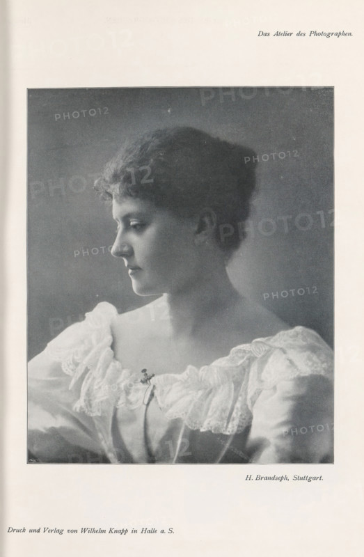 Portrait of an unknown woman, page, photomechanical print, inscription: 'Das Atelier des Photographen.', photographer: Hermann Brandseph, Stuttgart, cliché maker: Meisenbach, Riffarth & Cie., Berlin, printer (person): Wilhelm Knapp, Halle an der Saale, c. 1895 - in or before 1900, printer (person), Baden-Württemberg, Saxony-Anhalt, paper, autotypie, height 173 mm x width 140 mm, Print on third unnumbered page after page 28., historical persons - BB - woman, A black and white portrait of a young woman shown in profile looking left, with short wavy hair and a calm expression, wearing a light-colored dress with a wide lace-trimmed neckline and a small decorative clasp at the center, framed by a dark background and surrounded by a pale border.