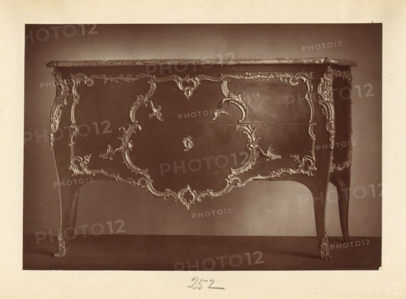 Chest of drawers in the style of Louis XV, from the collection of Christopher Beckett Denison, page, photomechanical print, number: '252', c. 1880 - in or before 1885, paper, Woodburytype, height 123 mm x width 178 mm, ornamental cupboard (wood and other phytogenic material), A photograph of the front and sides of a wooden table with a polished top and ornate carved decorations along its apron and legs, showing curved cabriole legs with carved floral and scroll motifs, a central carved panel with symmetrical scrollwork and foliage designs, decorative edging along the tabletop, and a smooth dark background behind the table.
