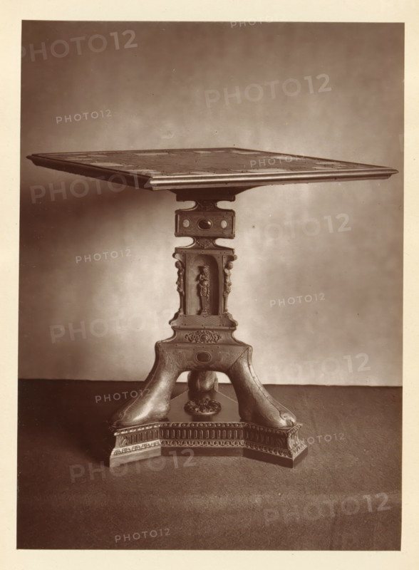 Table in front of a chessboard, from the collection of Christopher Beckett Denison, page, photomechanical print, number: '813', c. 1880 - in or before 1885, paper, Woodburytype, height 171 mm x width 125 mm, Print on fifth unnumbered page after page 80., chess, table (wood and other phytogenic material), A sepia-toned photograph of a decorative wooden table showing the square tabletop with a molded edge and a patterned surface, supported by a carved central pedestal. The pedestal features a rectangular section with rounded cutouts and ornamental oval insets, below which is a recessed niche containing a standing carved figure flanked by vertical decorative elements. The pedestal continues into a sculpted base with curved legs that sweep outward and rest on a molded plinth decorated with carved frieze patterns and a central floral medallion; the surface beneath the table appears to be a textured floor and a plain wall background.