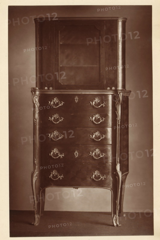 Chest of drawers in the style of Louis XV, from the collection of Christopher Beckett Denison, page, photomechanical print, number: '535', c. 1880 - in or before 1885, paper, Woodburytype, height 195 mm x width 121 mm, ornamental cupboard (wood and other phytogenic material), A tall wooden cabinet with an upper display section featuring a glass-fronted door and interior shelf sits atop a lower chest of drawers. The chest front shows ornate metal drawer pulls and escutcheons arranged symmetrically, with decorative carved appliques at the corners and along the vertical edges. The piece stands on curved, cabriole legs with carved detailing that continues up the sides. The wood surface has a polished finish with visible grain and subtle reflections, and the photograph is in sepia tones against a plain backdrop and floor.