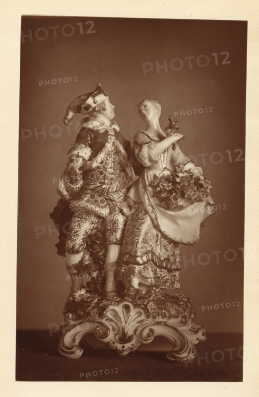 Sculpture of a personification of summer and winter, from the collection of Christopher Beckett Denison, page, photomechanical print, number: '3101', c. 1880 - in or before 1885, paper, Woodburytype, height 193 mm x width 121 mm, piece of sculpture, reproduction of a piece of sculpture, summer, 'Aestas', 'Estate' (Ripa), winter, 'Hyems', 'Inverno' (Ripa), A sepia-toned photograph of a porcelain figurine depicting a man and a woman standing close together on an ornate base, the man wearing a pointed hat and patterned clothing with a cloak draped over his shoulders and the woman in a flowing dress holding a small bouquet while lifting part of her skirt which is filled with flowers, both figures turned slightly toward each other, with detailed floral and scrollwork decoration on the base and textured patterns on the garments.