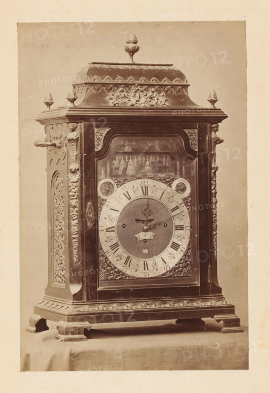 Clock, photograph, page, c. 1874 - in or before 1879, photographer, paper, albumen print, height 144 mm x width 95 mm, A vertically oriented photograph of an ornate wooden clock case standing on a draped surface, showing the upper decorative crest with a finial on top flanked by smaller finials, carved floral and foliate motifs along the cornice and sides, and a central glazed door revealing an inner painted scene above the clock face; the round clock dial has an inner circular plate with ornate hands and an outer ring marked with hour indicators and decorative spandrels, while the surrounding wood surfaces feature carved pilasters, applied scrollwork, and molded base with carved apron and bracket feet.