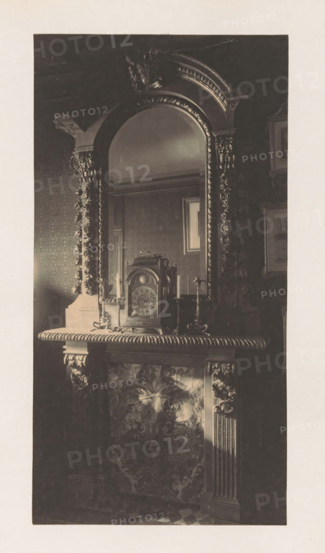 Chimney with clock in the house of Antonin Terme, photograph, page, Liège, c. 1880 - in or before 1885, photographer, paper, platinum process, height 212 mm x width 112 mm, clock, art collection, art gallery, chimney-piece, mantel piece, A dark photograph of an ornate mantel and mirror with heavy carved decoration framing an arched mirror, a decorative clock sitting on the mantel flanked by two candlesticks, patterned wallpaper and framed pictures visible in the reflected and surrounding walls, and sculpted floral and acanthus motifs on the mantel and pilasters beneath the shelf.