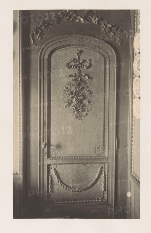 Wooden decorated door in the house of Antonin Terme, photograph, page, Liège, c. 1880 - in or before 1885, photographer, paper, platinum process, height 215 mm x width 132 mm, door, art collection, art gallery, sculpture (relief, sculpture), A tall wooden panel with an arched top is shown, featuring carved decorative motifs including a bow with hanging foliage near the upper center, ornate garlands draped across a lower recessed panel, and carved floral rosettes at the corners of a rectangular frame; the panel displays textured wood grain and raised moulding along its borders, with portions of adjacent wall and a series of decorative plates visible along the right edge.