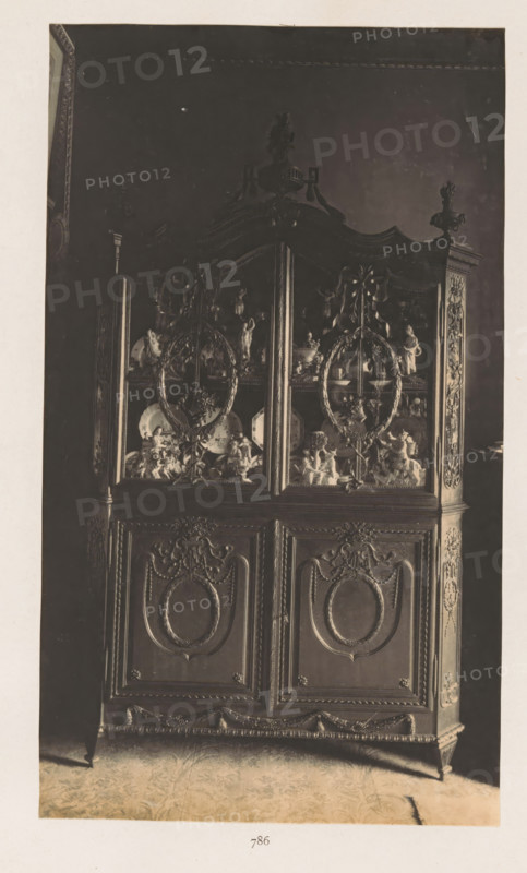 Cupboard in the house of Antonin Terme with statues and plates, photograph, page, Liège, c. 1880 - in or before 1885, photographer, paper, platinum process, height 221 mm x width 130 mm, 'Kunstschrank', art collection, art gallery, other rooms, A tall ornate wooden cabinet with carved panels and decorative molding fills the frame, its upper section featuring glass doors with intricate wreath-like carvings and visible porcelain plates and figurines inside, while the lower section displays raised oval medallions surrounded by carved garlands and scrollwork, all set against a dark background and standing on a patterned floor.