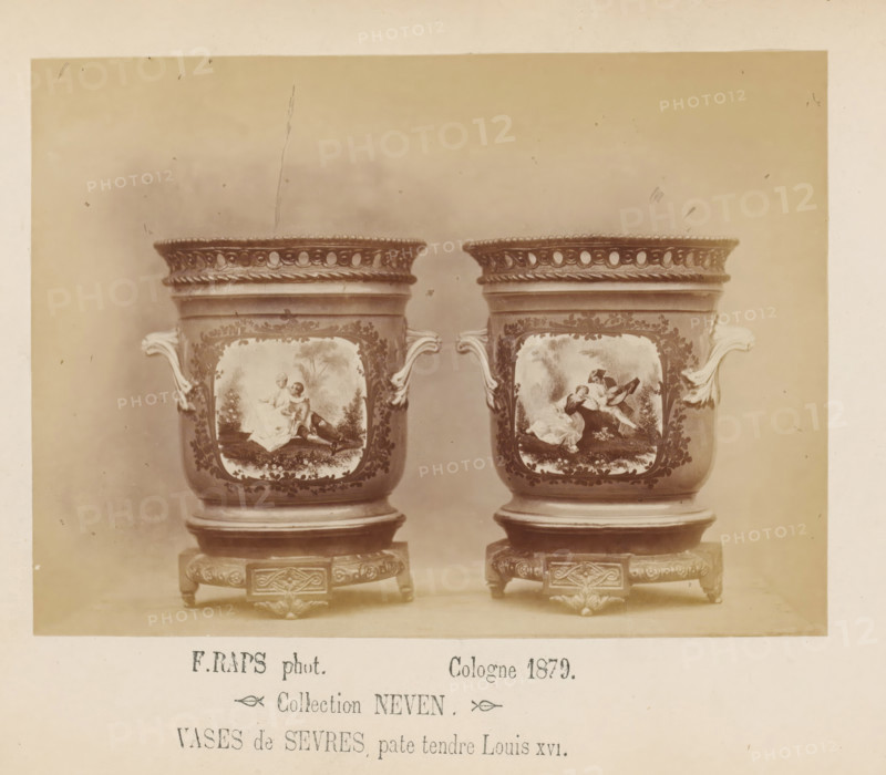 Two Sèvres vases of soft porcelain, photograph, page, inscription: 'Collection NEVEN, Vases de Sevres, pate tendre Louis XVI.', photographer: Friedrich Raps, Cologne, 1879, North Rhine-Westphalia, paper, albumen print, height 105 mm x width 142 mm, vase, ornament, A sepia-toned photograph showing two ornate ceramic vases placed side by side on decorative square stands, each vase featuring scroll-shaped handles and a decorative openwork rim, painted panels on their fronts depicting scenes with figures in classical dress set in landscapes, floral and foliate ornament surrounding the panels, and carved detailing on the wooden or metal stands beneath them.