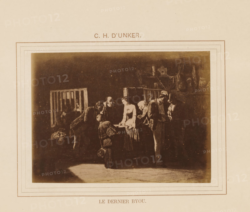 Photo reproduction of Le dernier byou by Carl Henrik D'Unker, Le dernier byou, photograph, reproduction, page, inscription: 'CH D'Unker', after painting by Carl Henrik D'Unker, c. 1878 - in or before 1883, Carl Henrik D'Unker, photographer, paper, albumen print, albumendruk, height 96 mm x width 131 mm, painting, drawing and the graphic arts, A sepia-toned print mounted on a pale page shows a dimly lit indoor scene with a group of people gathered around a central table. The group includes figures bending and standing, some wearing simple period clothing and one woman in a lighter dress leaning toward the table. A small child stands in the foreground holding a round object. The background contains wooden railings and indistinct interior elements, with darker shadows framing the scene and a lighter patch of floor in front of the group. The print is surrounded by decorative rectangular borders on the page.