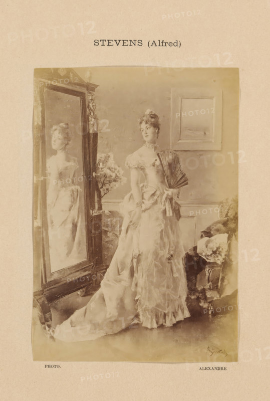 Stevens (Alfred), Photo reproduction of Jeune femme à sa toilette by Alfred Stevens, photograph, reproduction, page, photographer: Alexandre, after painting by Alfred Stevens, c. 1888 - in or before 1893, paper, albumen print, albumendruk, height 149 mm x width 105 mm, adolescent, young woman, maiden, looking in the mirror, contemplation of own beauty, painting, drawing and the graphic arts, A sepia-toned portrait of a woman in an elaborate gown standing beside a tall ornate mirror, the mirror reflecting her profile; she holds a folded fan at her side and wears a ruffled bodice with off-the-shoulder sleeves, and her hair is styled up with decorative accents; a framed picture hangs on the wall behind her and a cushioned chair with a patterned fabric sits to her side, while a small floral arrangement is visible near the mirror.