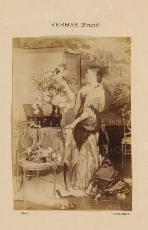 Photo reproduction of Le bouquet by Frans Verhas, Verhaz (Franz), photograph, reproduction, page, photographer: Alexandre, after painting by Frans Verhas, c. 1888 - in or before 1893, paper, albumen print, albumendruk, height 147 mm x width 101 mm, flowers, adolescent, young woman, maiden, painting, drawing and the graphic arts, A vertically oriented sepia photographic print shows a young woman standing beside a small table draped with a patterned cloth, reaching toward a large vase of flowers placed on the table; she wears a layered dress with a shiny skirt and a lace shawl over her shoulders, her hair pulled back in a braided style, and one hand rests on the table while the other holds a flower. The scene includes ornate chairs with carved details, a small framed item leaning on the table, scattered blossoms on the chair seat and floor, and a decorative folding screen or painted backdrop behind the woman depicting trees and architectural forms. The image is mounted on a textured ligh...