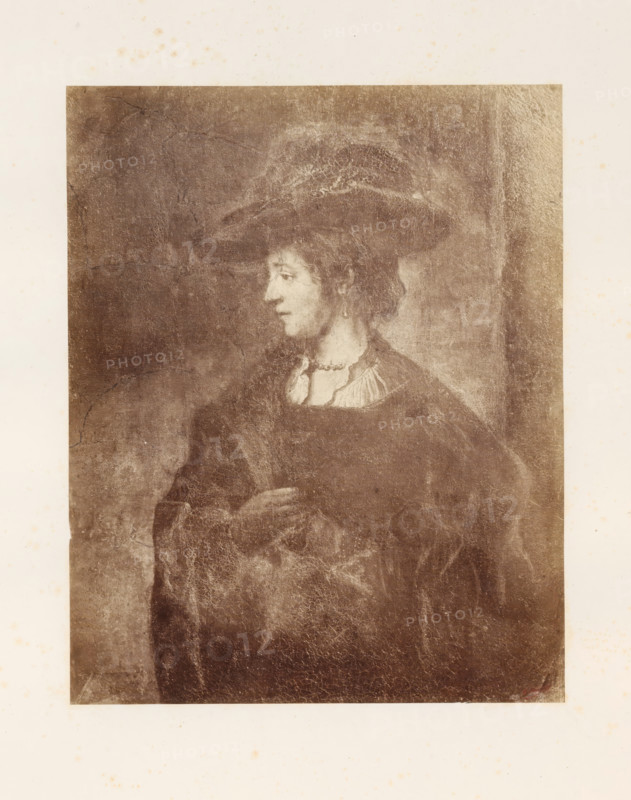 Photo reproduction of Saskia van Uylenburgh by Rembrandt van Rijn, photograph, reproduction, page, photographer: Edmond Fierlants, after painting by Rembrandt van Rijn, publisher: C. Muquardt, Brussels, c. 1856 - in or before c. 1861, paper, albumen print, height 352 mm x width 281 mm, Person, Saskia van Uylenburgh, historical persons - BB - woman, A sepia-toned portrait of a person shown from the waist up facing left, wearing a wide-brimmed hat and a dark robe with a lighter collar, with an earring visible on the left ear and one hand resting near the chest; the image shows visible crackling and aged texture across the surface and a lighter border surrounding the portrait.