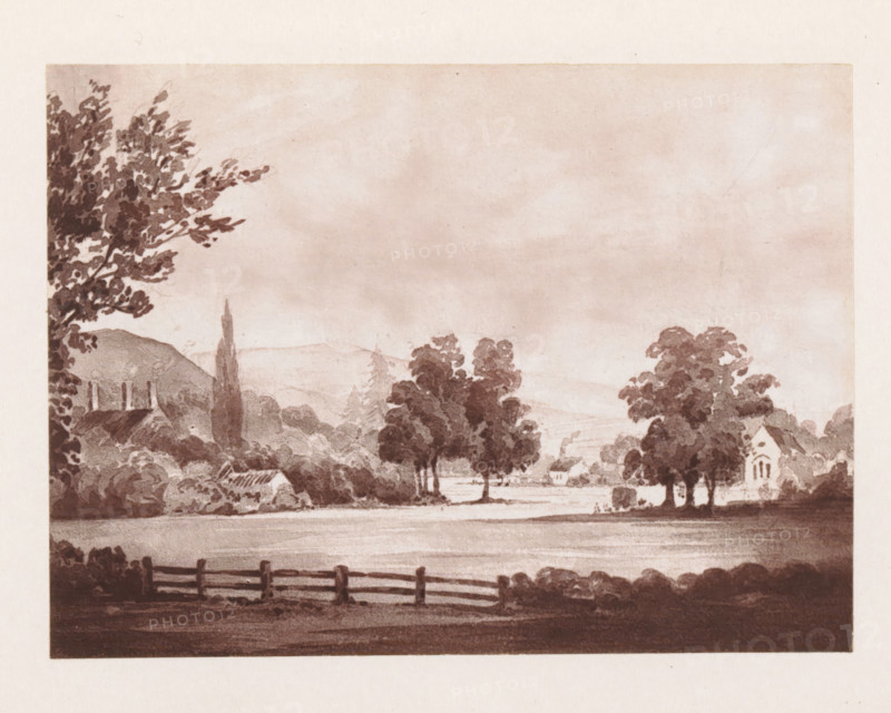 Photo reproduction of a painting depicting a view of Alton Barnes, reproduction, page, photomechanical print, cliché maker: Woodbury Permanent Photographic Printing Company, after painting by Miss Clinton, Alton (Wiltshire), c. 1866 - in or before 1876, photographer, Wiltshire, paper, Woodburytype, height 84 mm x width 114 mm, city-view in general, 'veduta', A sepia-toned landscape showing a wide foreground with a wooden fence along the bottom edge, a body of water or flat field behind it, and clusters of trees of varying shapes and sizes rising toward the middle ground; on the left a tall tree with dense foliage frames the scene, a low roofline and chimneys are partly visible behind shrubbery, and a slender tall tree stands near rolling hills; on the right a small building with arched windows sits among trees, while distant hills and a cloudy sky fill the upper portion of the image.