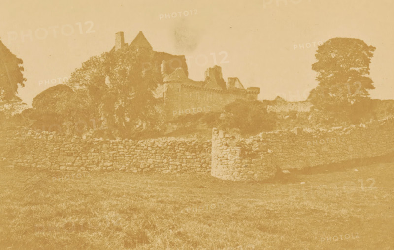 Craigmillar, View of Craigmillar Castle, photograph, page, number: 'Pl. 2.', photographer: Vincent Brooks, Day & Son, Edinburgh, c. 1864 - in or before 1874, paper, albumen print, height 77 mm x width 119 mm, castle, Craigmillar Castle, A faded sepia-toned photograph shows an old stone wall running horizontally across a grassy foreground, with a low stone pillar interrupting the wall near the center. Behind the wall, there are dense trees and foliage partially obscuring the outline of a ruined stone building with crenellated walls and angled rooflines. A large, rounded tree stands to the right of the building, and additional trees and vegetation frame the left side. The sky above is pale and mostly empty, and the image has a worn, aged appearance with uneven toning.