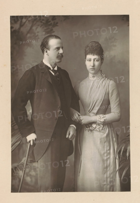 The Duke and Duchess of Fife, Portrait of Alexander Duff and his wife Louise Victoria Alexandra Dagmar of Saxe-Coburg and Gotha, page, photomechanical print, inscription: '57 & 61, Elbury Street, London.', photographer: Downey, William & Daniel, London, clichémaker: anonymous, c. 1880 - in or before 1890, paper, Woodburytype, Woodburytypie, height 139 mm x width 94 mm, Person, Louise Victoria Alexandra Dagmar (Princess of the United Kingdom), Alexander William George Duff (Duke of Fife), historical persons, historical persons - BB - woman, A sepia-toned portrait shows a standing man and woman positioned close together against a plain backdrop with faint foliage shadows; the man wears a dark three-piece suit with a high-collared shirt and tie, has short hair and a mustache, and holds a cane in one hand while his other hand rests near his waist; the woman wears a light-colored, high-necked dress with pleated and ruffled details at the bodice and cuffs, has hair styled up, and clasps her ...