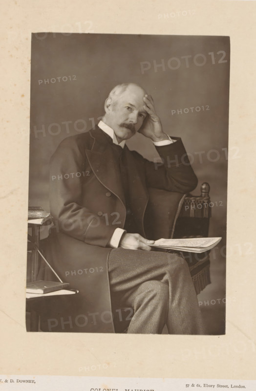 Portrait of John Frederick Maurice, Colonel Maurice, page, photomechanical print, inscription: '57 & 61, Elbury Street, London.', photographer: Downey, William & Daniel, London, cliché maker: anonymous, c. 1880 - in or before 1890, paper, Woodburytype, height 141 mm x width 94 mm, Person, John Frederick Maurice, commander-in-chief, general, marshal, A sepia portrait of an older man seated in a chair, shown from head to mid-thigh, wearing a dark formal coat with a high collar and light shirt cuffs, with one hand resting on his forehead and the other holding a folded newspaper on his knee; he has a mustache and short hair, and the chair beside him has a fringed arm and a rounded finial, with a small side table holding stacked books partially visible.