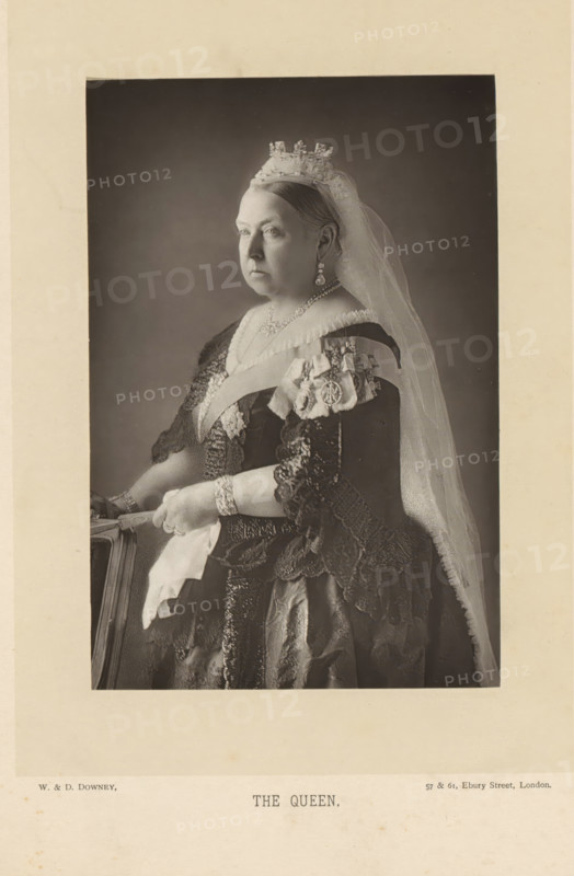 The Queen, Portrait of Victoria of the United Kingdom, page, photomechanical print, inscription: '57 & 61, Elbury Street, London.', photographer: Downey, William & Daniel, London, clichémaker: anonymous, c. 1882 - in or before 1892, paper, Woodburytype, Woodburytypie, height 140 mm x width 93 mm, Person, Victoria (Queen of the United Kingdom and Empress of India), ruler, sovereign - BB - female ruler, A sepia-toned portrait of an older woman shown from the waist up, seated in profile facing left, wearing a light jeweled crown and a long translucent veil that falls over her shoulder; she has a solemn expression and her hair is pulled back, and she is dressed in an ornate dark gown with lace sleeves and layered fabrics, adorned with decorative sashes, a brooch and hanging earrings, and she rests her gloved hands on the back of a chair while wearing bracelets and holding a folded handkerchief.