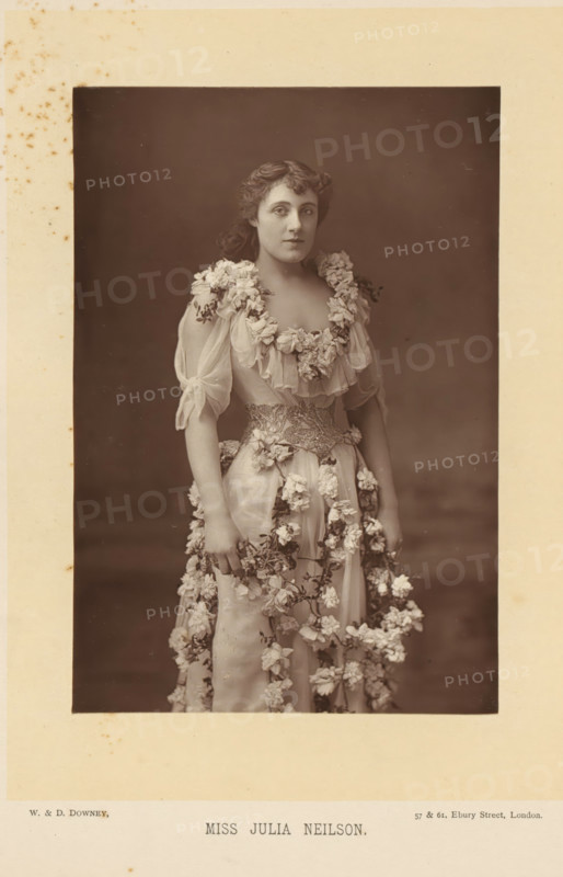 Portrait of Julia Neilson in a dress with flowers, Miss Julia Neilson, page, photomechanical print, inscription: '57 & 61, Elbury Street, London.', photographer: Downey, William & Daniel, London, cliché maker: anonymous, c. 1882 - in or before 1892, paper, Woodburytype, height 140 mm x width 95 mm, Person, Julia Neilson, portrait of actor, actress, A sepia studio portrait of a young woman standing and facing forward, wearing a light, flowing gown decorated with clusters of flowers around the neckline, sleeves, waist, and cascading down the skirt; her hair is wavy and pulled back from her face, she has a neutral facial expression and is looking slightly to the side, her bare arms are visible and her hands rest at her sides holding part of the floral garlands, the background is plain and dark, and the photograph is mounted on a light-colored card with some small spots and foxing visible around the edges.