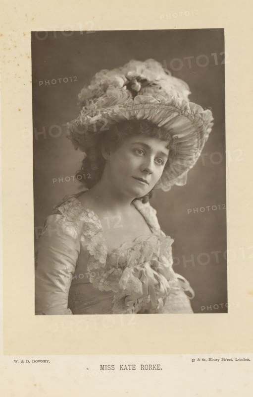 Miss Kate Rorke, Portrait of Kate Rorke, page, photomechanical print, inscription: '57 & 61, Elbury Street, London.', photographer: Downey, William & Daniel, London, cliché maker: anonymous, c. 1882 - in or before 1892, paper, Woodburytype, height 140 mm x width 95 mm, Person, Kate Rorke, portrait of actor, actress, A sepia portrait of a woman shown from the chest up wearing a wide, ruffled hat decorated with layered fabric and flowers, with curly hair visible beneath the brim; she has a soft, neutral facial expression and is turned slightly to one side; her dress has lace trim around the neckline and bodice and features bows and gathered fabric; the background is plain and evenly shaded and the portrait is mounted on a bordered card with visible aging spots.