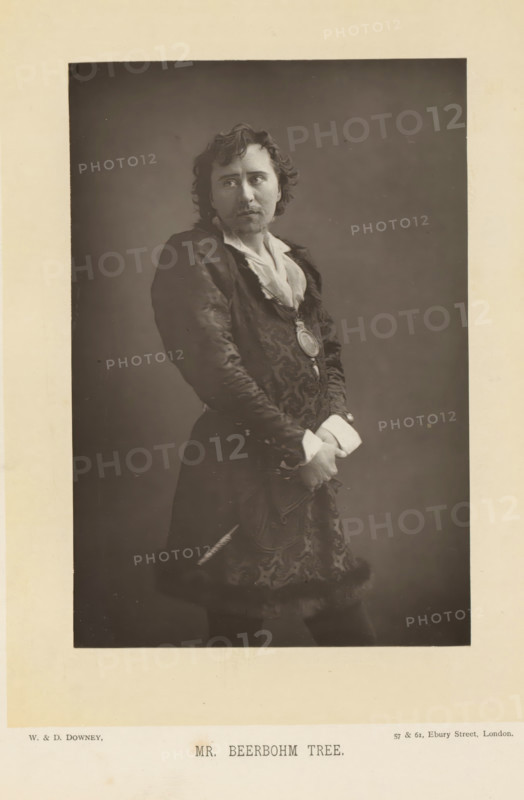 Mr. Beerbohm Tree, Portrait of Herbert Beerbohm Tree, page, photomechanical print, inscription: '57 & 61, Elbury Street, London.', photographer: Downey, William & Daniel, London, cliché maker: anonymous, c. 1882 - in or before 1892, clichémaker, paper, Woodburytype, Woodburytypepie, height 141 mm x width 95 mm, Person, Herbert Beerbohm Tree, portrait of actor, actress, stage costume, A sepia-toned portrait of a man shown from head to mid-thigh against a plain darker background, wearing a patterned dark coat with fur trim at the hem and a light-colored shirt with an open collar, his hair wavy and shoulder-length, a mustache and small goatee on his face, a round medallion suspended on a ribbon at his chest, his hands clasped in front at waist level with visible light-colored cuffs, a slender object tucked at his side, and a neutral expression with his gaze directed slightly to one side.