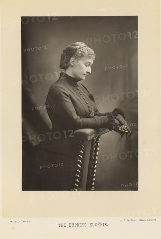 The empress Eugénie, Portrait of Eugénie de Montijo, page, photomechanical print, inscription: '57 & 61, Elbury Street, London.', photographer: Downey, William & Daniel, London, cliché maker: anonymous, c. 1882 - in or before 1892, paper, Woodburytype, height 140 mm x width 93 mm, Person, Eugénie (Empress of the French), ruler, sovereign - BB - female ruler, A sepia-toned portrait of a seated woman shown in profile looking downward, wearing a dark high-collared dress with a row of fabric-covered buttons down the front and decorative ruffled cuffs, her hair covered by a textured cap, hands resting on the padded arm of a turned-wood chair while holding a small cluster of flowers, a softly blurred backdrop behind her and a wide pale margin framing the photograph.