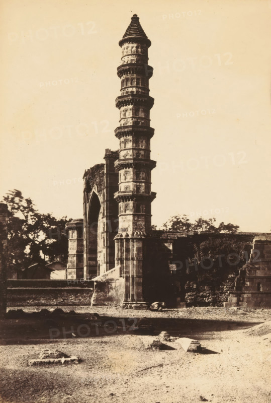 Mosque of Mahomed Ghous.- From the North, View of a minaret of the Mohammed Ghous Mosque in Ahmedabad, photograph, page, inscription: '2D2', number: '102.', photographer: Thomas Biggs, Ahmedabad (city), c. 1856 - in or before 1866, Gujarat, paper, albumen print, height 189 mm x width 137 mm, temple, shrine, Islam, Mohammedanism, Mohammed Ghous Mosque, A tall stone tower with multiple circular balconies rising above a partially ruined arched structure, surrounded by remnants of masonry and low walls, with patches of vegetation and trees visible behind and to the left of the buildings, and a foreground of bare ground scattered with small stone fragments.