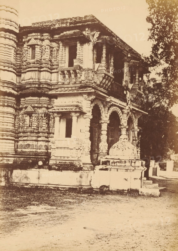 View of the Hutheesing Temple in Ahmedabad, Shet Huttising's Temple.- The Porch, photograph, page, number: '119.', photographer: Thomas Biggs, Ahmedabad (city), c. 1856 - in or before 1866, Thomas Biggs, photographer, Gujarat, paper, albumen print, height 197 mm x width 144 mm, places and objects of worship, Hinduism, Buddhism, Jainism, Hutheesing temple, A sepia-toned photograph of a carved stone building façade with ornate columns, balustrades, and arched openings, showing layered horizontal bands of decorative relief and sculpted figures on the walls; a small dome-like stone structure sits on a raised platform near the entrance steps, and trees are visible to the right, with a textured ground area in the foreground.
