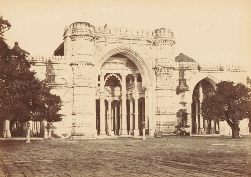 Façade of the Jama Masjid in Ahmedabad, The Jumma Mosque.- Nearer view, photograph, page, number: '15.', photographer: Thomas Biggs, Ahmedabad (city), c. 1856 - in or before 1866, Gujarat, paper, albumen print, height 136 mm x width 189 mm, temple, shrine, Islam, Mohammedanism, parts of church exterior and annexes: facade, Jama Masjid (Ahmedabad), A sepia-toned photograph of a large ornate stone gateway with a central pointed arch flanked by tall cylindrical columns with decorative horizontal bands, a series of slender columns and scalloped arches visible within the entrance, and carved architectural detailing along the top edge; trees frame both sides of the gateway, casting shadows on the facade and standing beside a paved courtyard in the foreground.