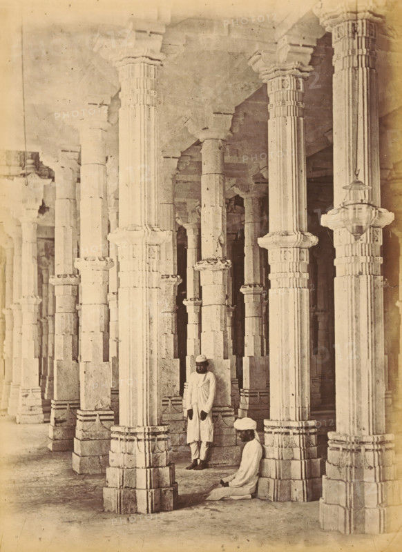 Interior of the Jama Masjid in Ahmedabad, The Jumma Mosque.- Pillars in the Interior, photograph, page, number: '18.', inscription: 'F2', photographer: Thomas Biggs, Ahmedabad (city), c. 1856 - in or before 1866, Gujarat, paper, albumen print, height 201 mm x width 148 mm, temple, shrine, Islam, Mohammedanism, column, pillar, architecture, Jama Masjid (Ahmedabad), A sepia-toned photograph showing a large interior space filled with tall carved stone pillars arranged in rows, each pillar featuring fluted shafts and ornate capitals and bases with layered stonework; two men in traditional light-colored clothing and turbans are among the pillars, one standing and leaning against a pillar and the other seated on the floor near a base; the floor appears smooth and worn, the ceiling is supported by the pillars with visible stone beams, and a hanging lantern is suspended from one pillar.