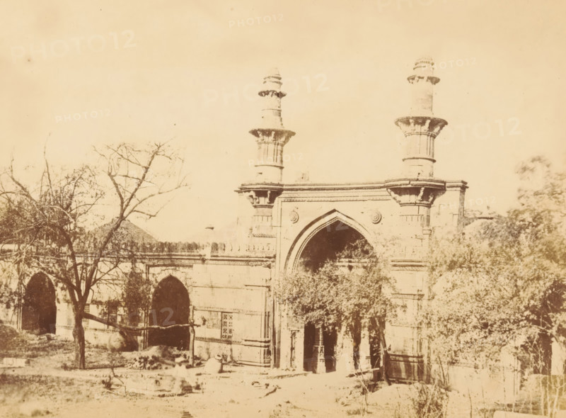 Ahmed Shah's Mosque, View of Ahmed Shah's Mosque in Ahmedabad, photograph, page, number: '2.', photographer: Thomas Biggs, Ahmedabad (city), c. 1856 - in or before 1866, Gujarat, paper, albumen print, height 136 mm x width 184 mm, temple, shrine, Islam, Mohammedanism, Ahmed Shah Mosque, A sepia-toned photograph showing a stone gateway with an arched central entrance flanked by decorative columns and two tall cylindrical towers rising above the structure, leafless trees and foliage partially obscuring the lower sections of the building, a series of smaller arches along the facade to the left, and scattered stones and ground-level debris in the foreground.