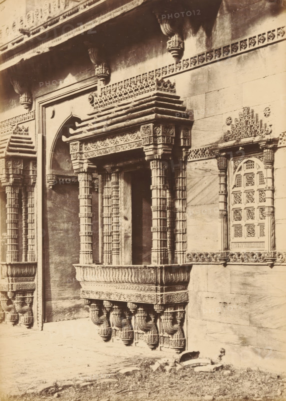 Façade of the Rani Rupamati Mosque in Ahmedabad, The Queen's Mosque in Mirzapoor.- The Northern Side-arch and Windows, photograph, page, number: '31.', photographer: Thomas Biggs, Ahmedabad (city), c. 1856 - in or before 1866, Thomas Biggs, photographer, Gujarat, paper, albumen print, height 188 mm x width 135 mm, temple, shrine, Islam, Mohammedanism, parts of church exterior and annexes: facade, Rani Rupamati Mosque, A sepia photograph of a stone building façade featuring ornate carved columns, brackets, and decorative ledges forming a balcony-like projection, with intricately patterned lattice window panels set into the wall; the carved elements include layered cornices, geometric and floral motifs, and sculpted supports beneath the projecting platform; a framed arched recess appears beside the central projection, and a patterned frieze runs horizontally across the upper wall; the ground in front shows rough stone and sparse grass.